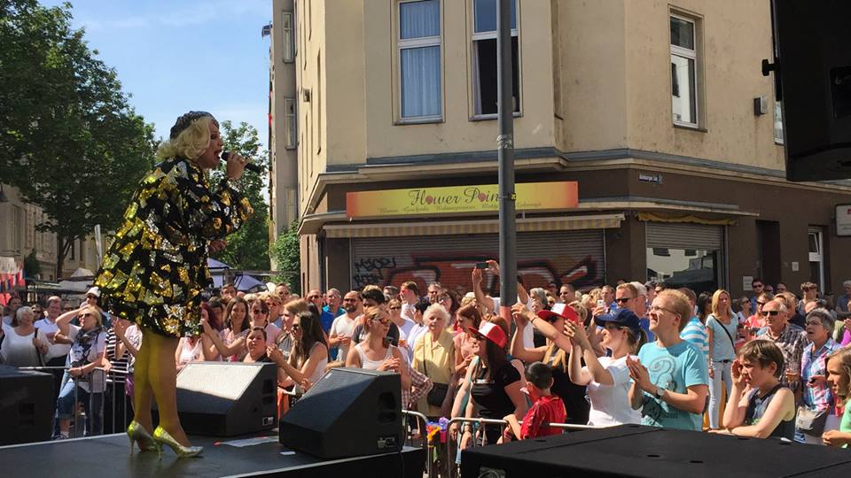 Eine Sängerin in einem gelb-schwarzen Kleid performt auf einer Bühne vor einer begeisterten Menschenmenge bei einer Veranstaltung im Freien.