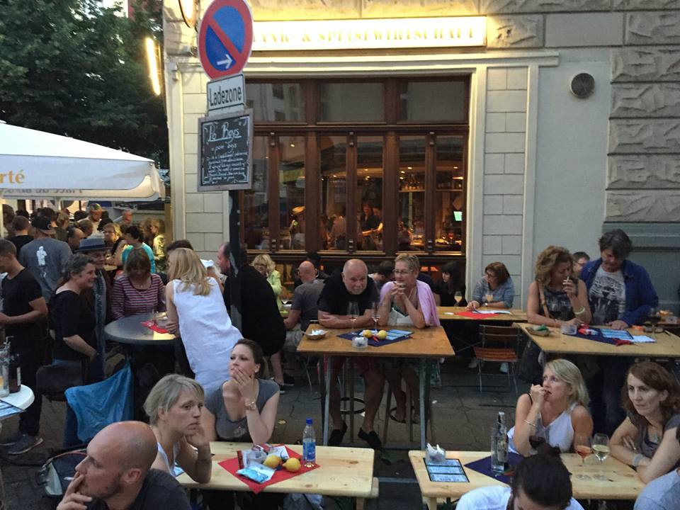 Gäste sitzen an Tischen vor einem Restaurant im Freien und genießen die abendliche Atmosphäre mit Gesprächen und Getränken unter einem