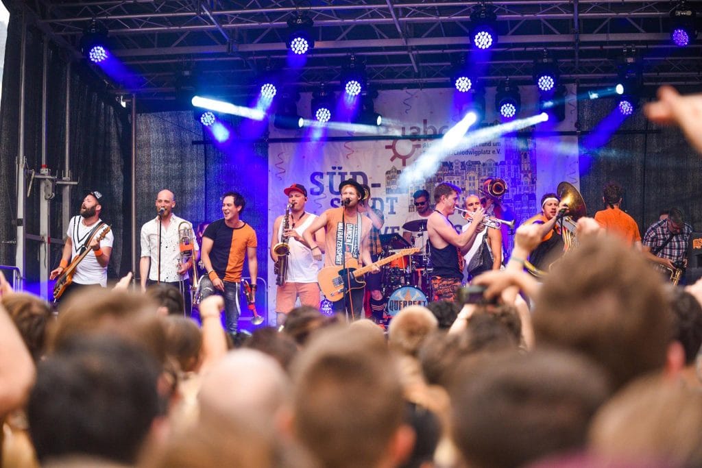 Eine Band spielt auf der Bühne des Südstadtfestes vor einem begeisterten Publikum, das dicht gedrängt vor der Bühne steht und die Musik feiert, während bunte Scheinwerfer die Szene in helles Licht tauchen.