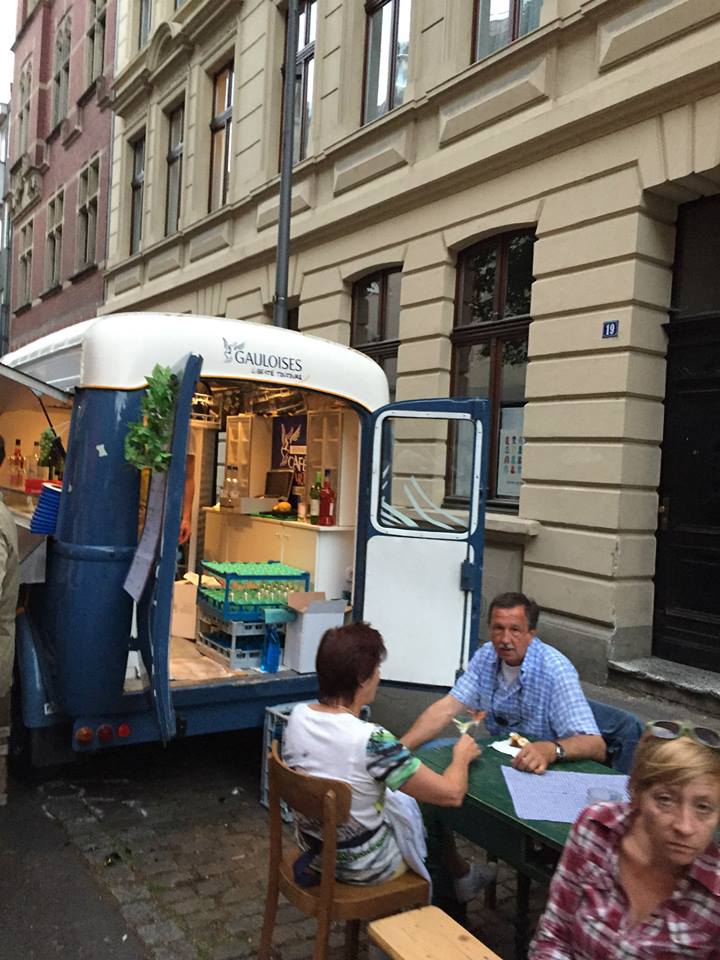 Besucher sitzen an Tischen vor einem umgebauten blauen Verkaufswagen, der als Bar dient, während sie Getränke genießen und sich unterhalten.