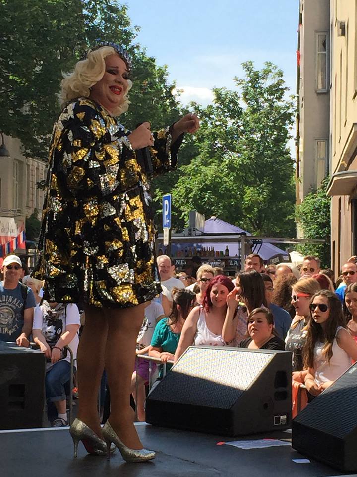 Eine Dragqueen steht auf einer Bühne und spricht in ein Mikrofon, umgeben von Zuschauern bei einer Veranstaltung im Freien.