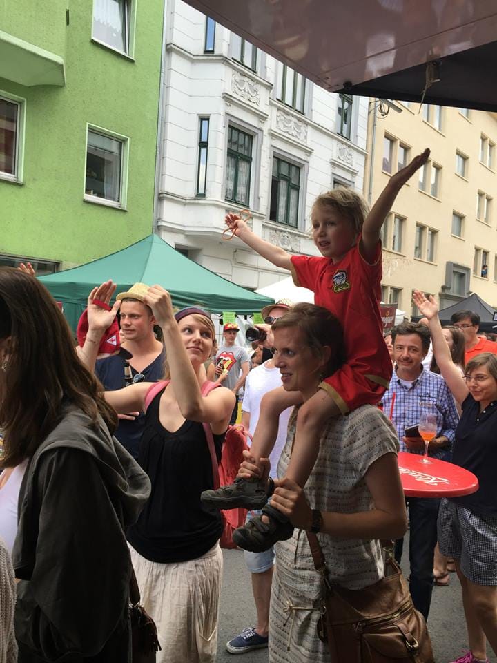 Besucher feiern auf einer belebten Straße, ein Kind sitzt auf den Schultern einer Frau und jubelt mit erhobenen Armen in