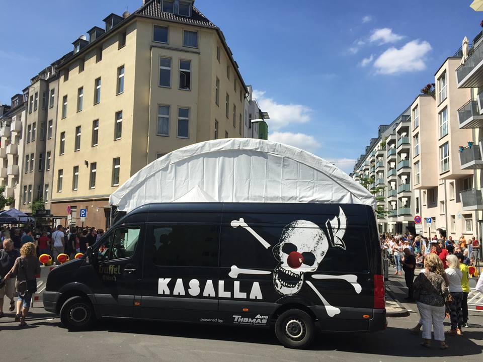 Ein schwarzer Van mit Totenkopf-Motiv und der Aufschrift Kasalla steht vor einer Menschenmenge und Gebäuden unter blauem Himmel.