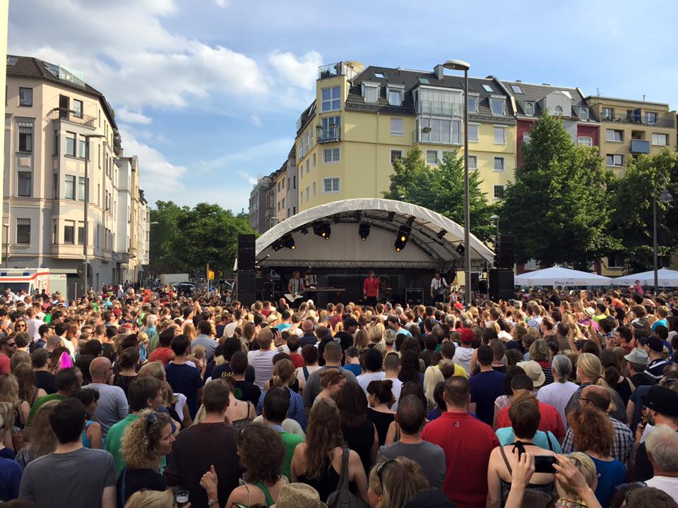 Besucher feiern beim Südstadtfest in Köln vor einer Bühne mit Musikern, umgeben von Wohnhäusern und Bäumen, während sie die sommerliche Atmosphäre genießen und die Musik der Live-Band im Freien erleben.