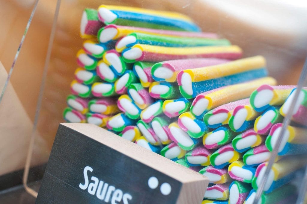 Süßigkeiten in Regenbogenfarben liegen gestapelt in einem durchsichtigen Behälter mit einem kleinen Schild, auf dem Saures geschrieben steht.