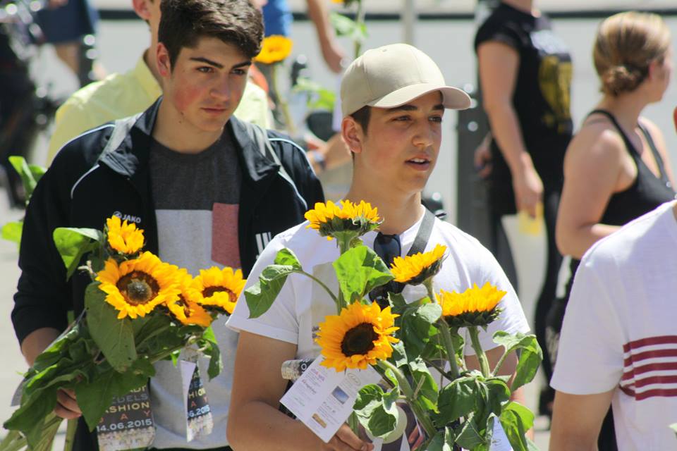 Zwei junge Männer tragen Sonnenblumen in der Hand und gehen aufmerksam durch eine Menschenmenge im Freien.