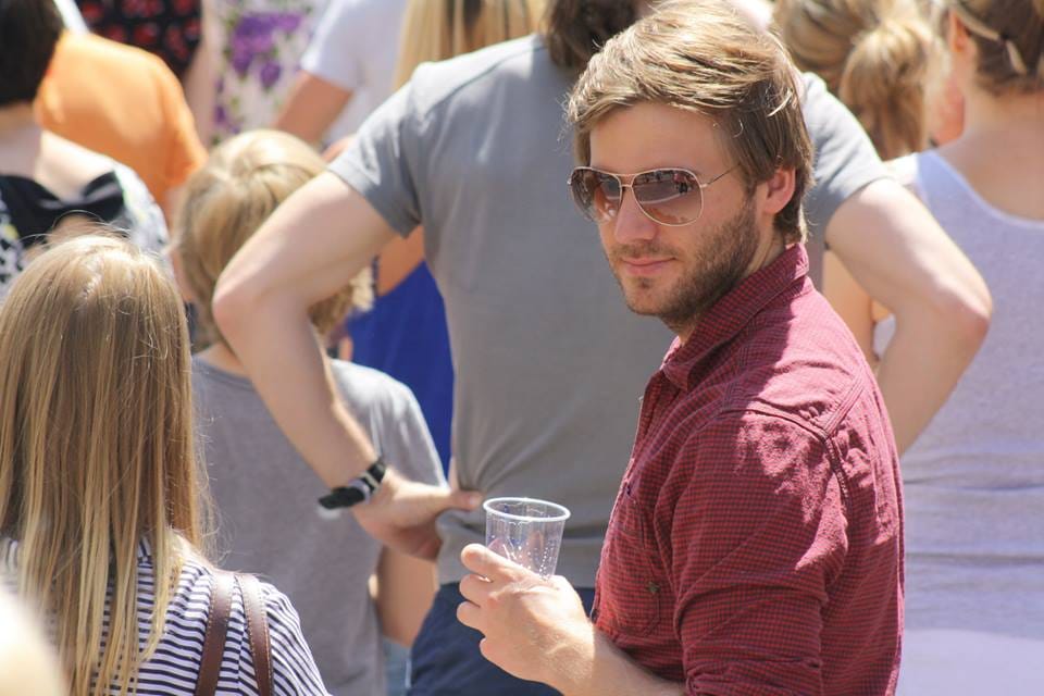 Ein Mann mit Sonnenbrille und rotem Hemd hält einen Plastikbecher in der Hand und schaut in die Kamera, umgeben von