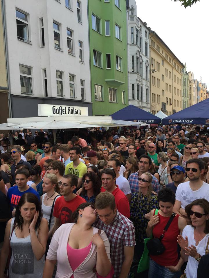 Besucher drängen sich an einem sonnigen Tag unter einem Bierstand, einige trinken und unterhalten sich, während im Hintergrund Wohnhäuser zu