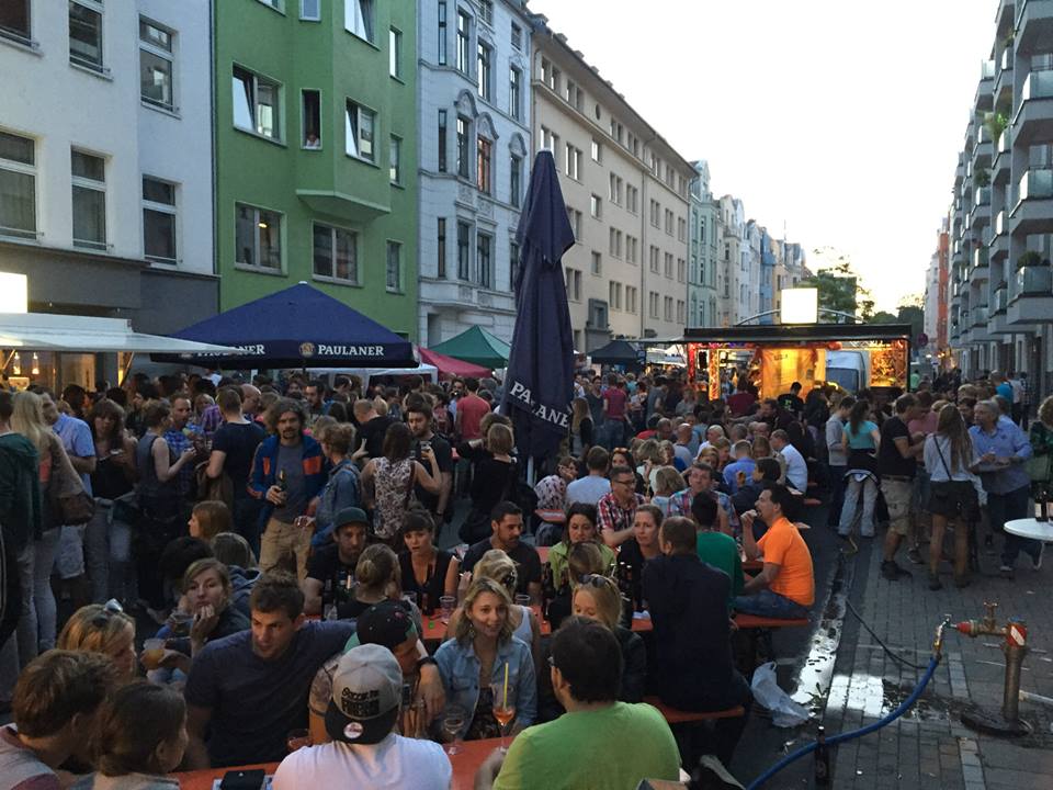 Besucher drängen sich auf einer belebten Straße mit Imbissständen und Zelten, umgeben von hohen Wohnhäusern, während sie Getränke genießen und