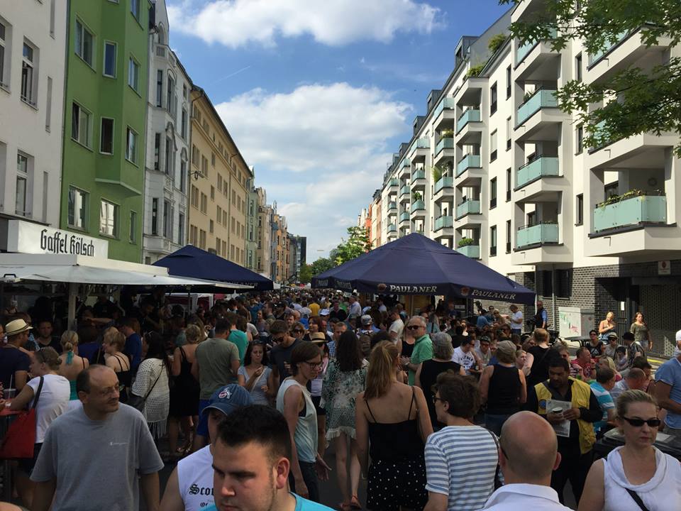 Besucher schlendern an einem sonnigen Tag durch eine belebte Straße mit Imbissständen und bunten Wohnhäusern, genießen die lebhafte Atmosphäre.