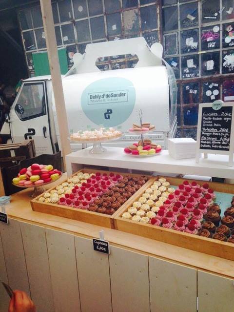 Ein Verkaufsstand mit einer Auswahl an bunten Macarons und kleinen Törtchen, präsentiert auf Holztabletts, mit einem weißen Lieferwagen im Hintergrund.