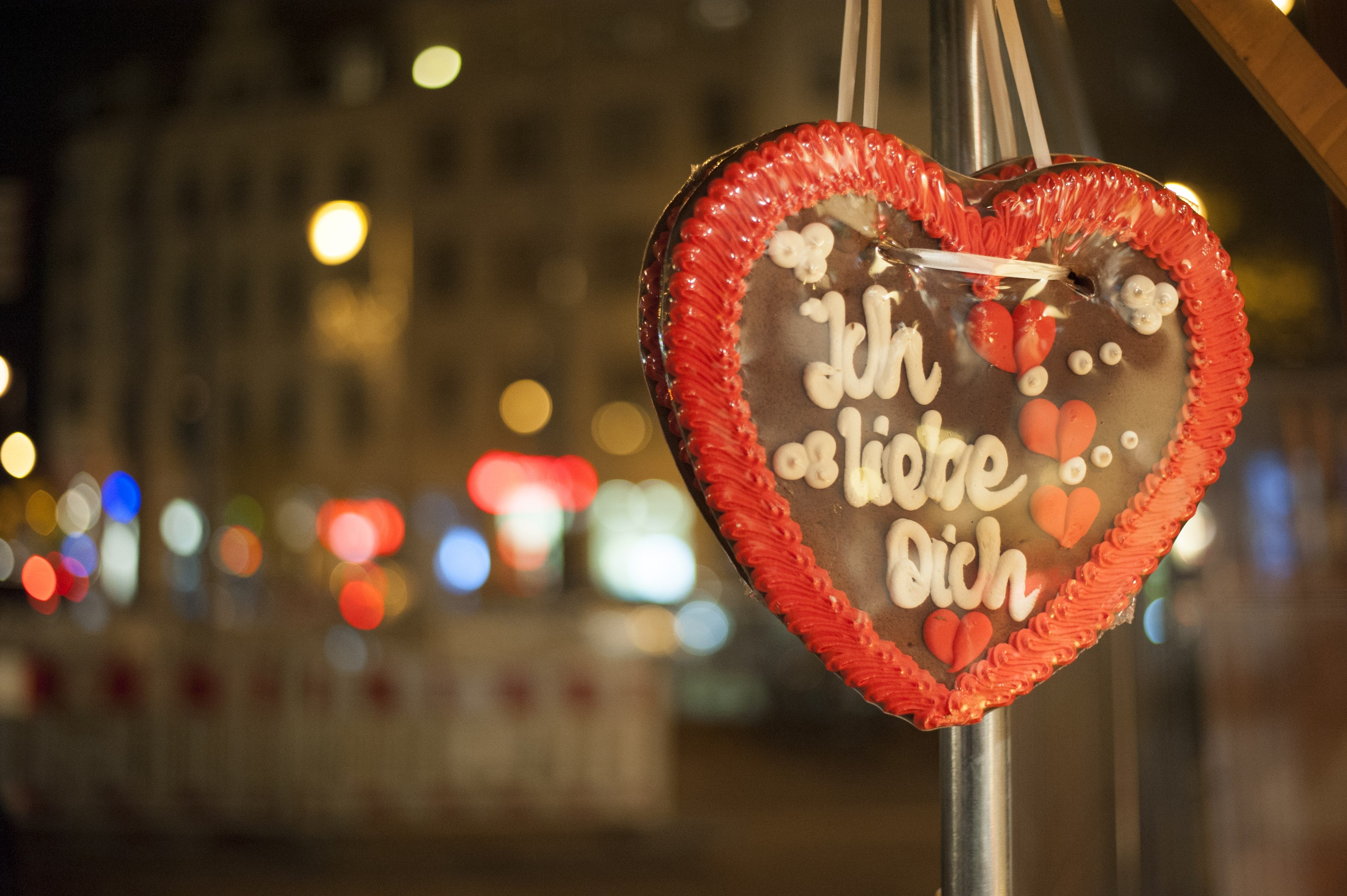 Ein Lebkuchenherz mit der Aufschrift Ich liebe dich hängt an einem Stand, im Hintergrund verschwommene Lichter einer abendlichen Veranstaltung.