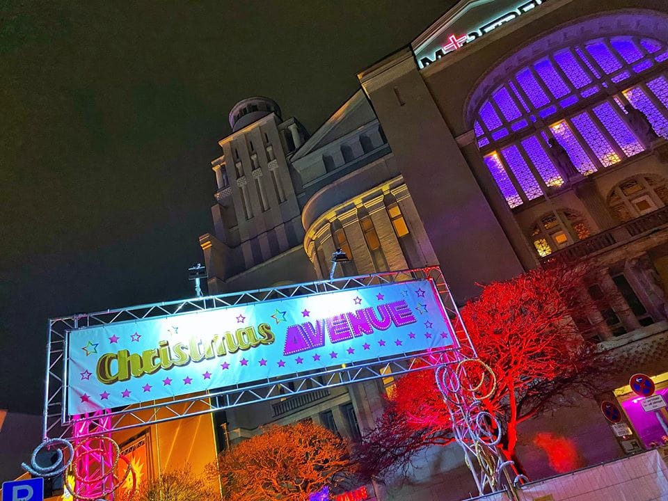 Einladendes Schild mit der Aufschrift Christmas Avenue vor einem beleuchteten Gebäude am Nollendorfplatz in Berlin, das mit seiner festlichen Dekoration und dem bunten Lichtspiel eine weihnachtliche Atmosphäre schafft und Besucher anzieht.
