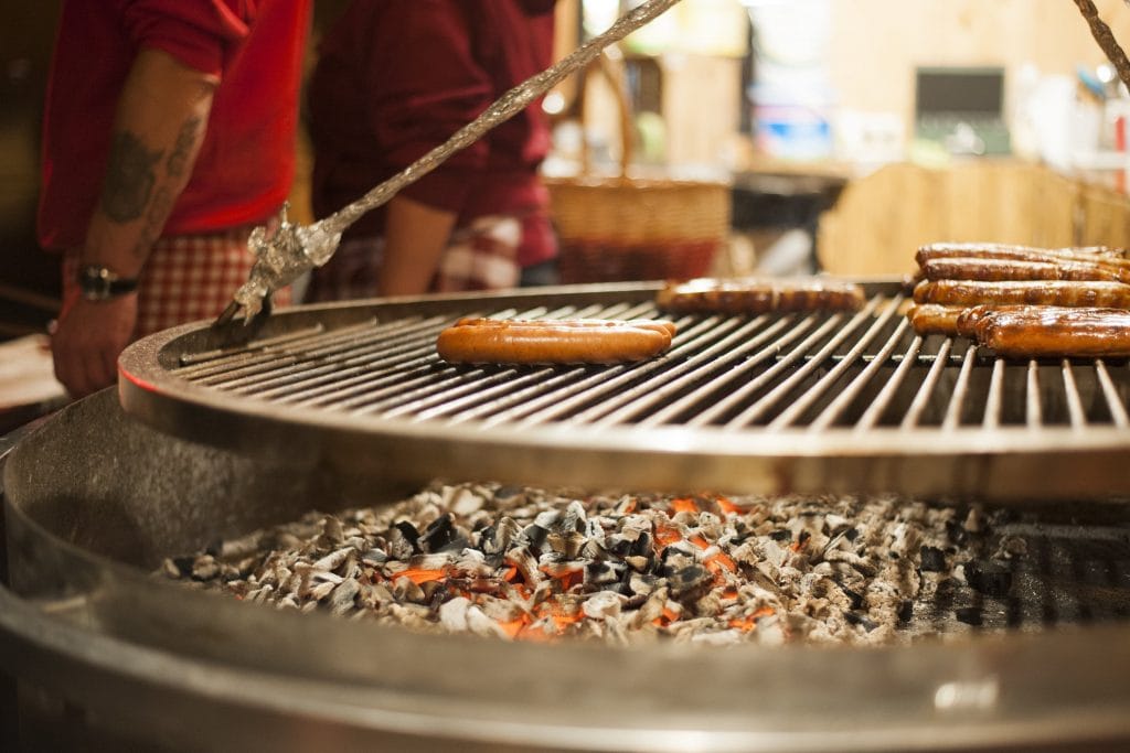 Ein Koch grillt auf dem VeedelsAdvent Würstchen auf einem großen Holzkohlegrill, während er mit einer Zange die Würste wendet, um sie gleichmäßig zu bräunen. Die glühende Holzkohle spendet Wärme und Licht, während die Besucher des Weihnachtsmarktes hungrig auf die frisch gegrillten Würste warten.