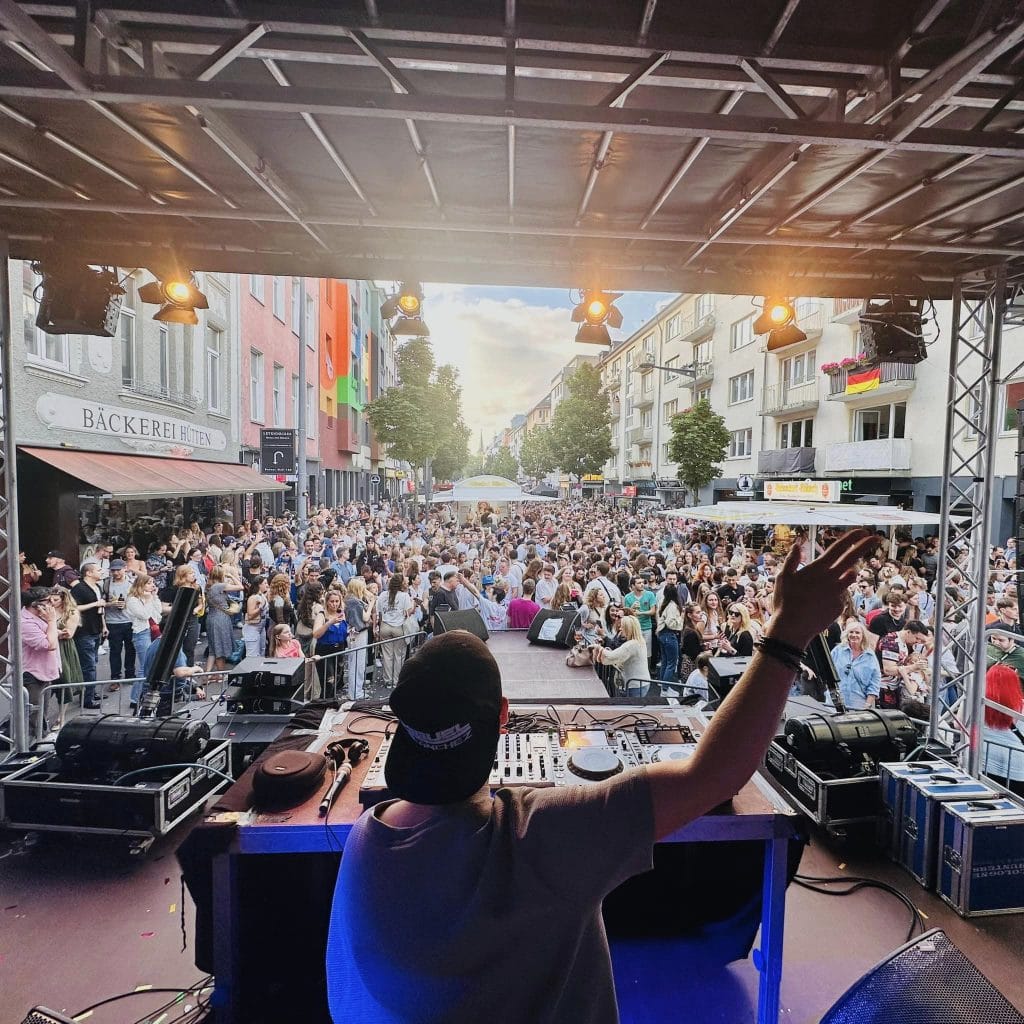 Ein DJ legt beim Südstadtfest in Köln auf einer Bühne vor einem großen Publikum auf, das sich in der belebten Straße versammelt hat und die Musik genießt, während die Lichter der Bühne die Szene erhellen.