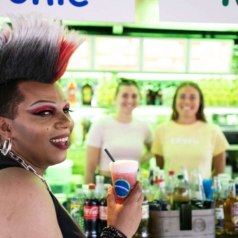 Eine Dragqueen mit extravaganten Haaren und Make-up hält einen Cocktail vor einem Getränkestand mit zwei lächelnden Frauen im Hintergrund.