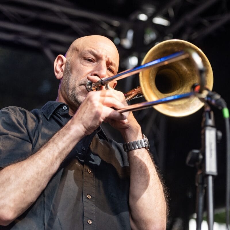 Ein Posaunist mit Hemd und Armbanduhr spielt engagiert auf seinem Instrument bei einem Konzert.