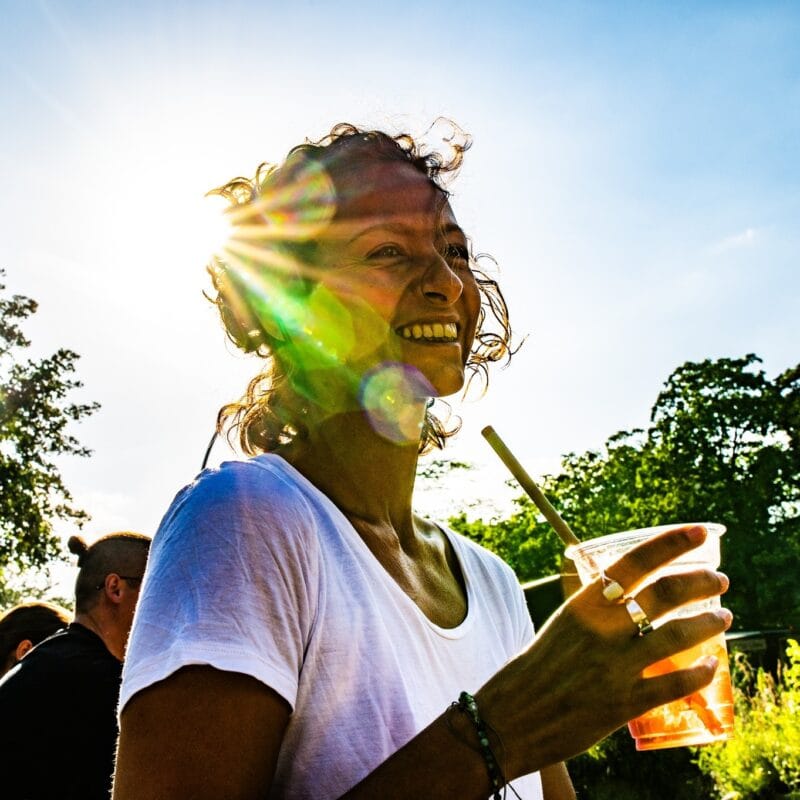 Eine lachende Frau im Gegenlicht genießt einen erfrischenden Cocktail im Freien, umgeben von Bäumen und anderen Event-Besuchern.