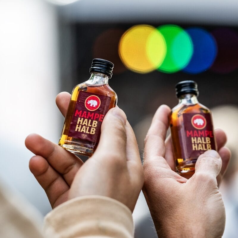 Zwei Hände halten Mampes Halb und Halb Miniaturflaschen hoch, im Hintergrund sind verschwommene Lichter zu sehen.