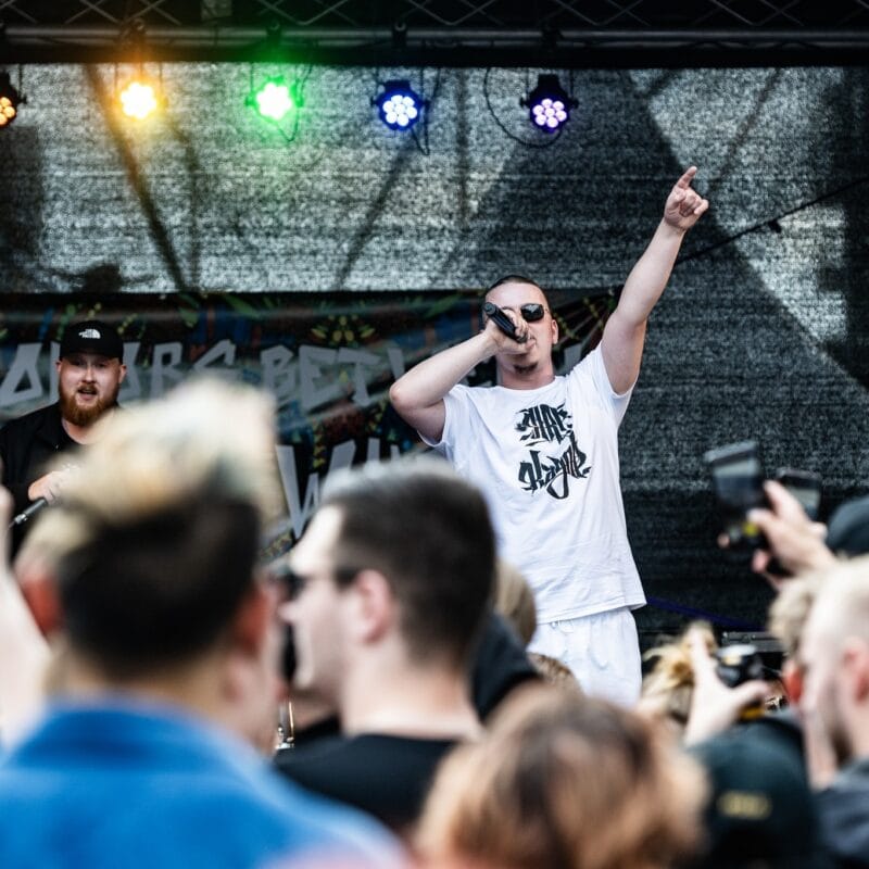 Ein Rapper in weißem T-Shirt performt auf einer Bühne vor einem begeisterten Publikum, das mit Handys filmt.