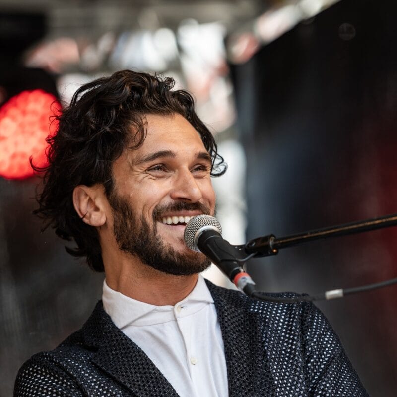 Ein lächelnder, bärtiger Sänger im dunklen Sakko singt bei einem Auftritt ins Mikrofon.