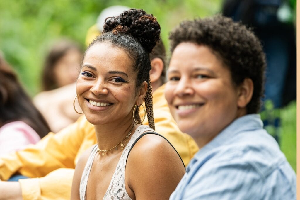 Zwei Frauen lächeln in die Kamera, eine mit Dutt und die andere mit kurzen Haaren.