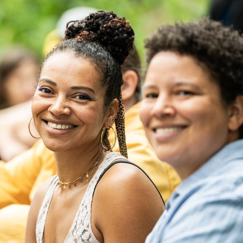 Zwei Frauen lächeln in die Kamera, eine mit Dutt und die andere mit kurzen Haaren.