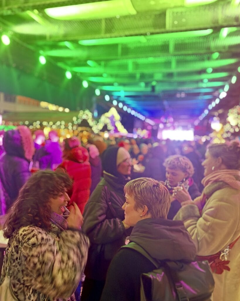 Zwei junge Frauen unterhalten sich auf einem Weihnachtsmarkt, während im Hintergrund festliche Beleuchtung und weitere Besucher zu sehen sind.