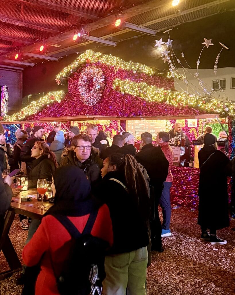 Besucher stehen vor einem festlich geschmückten Stand auf dem Weihnachtsmarkt und genießen die weihnachtliche Atmosphäre mit Beleuchtung.