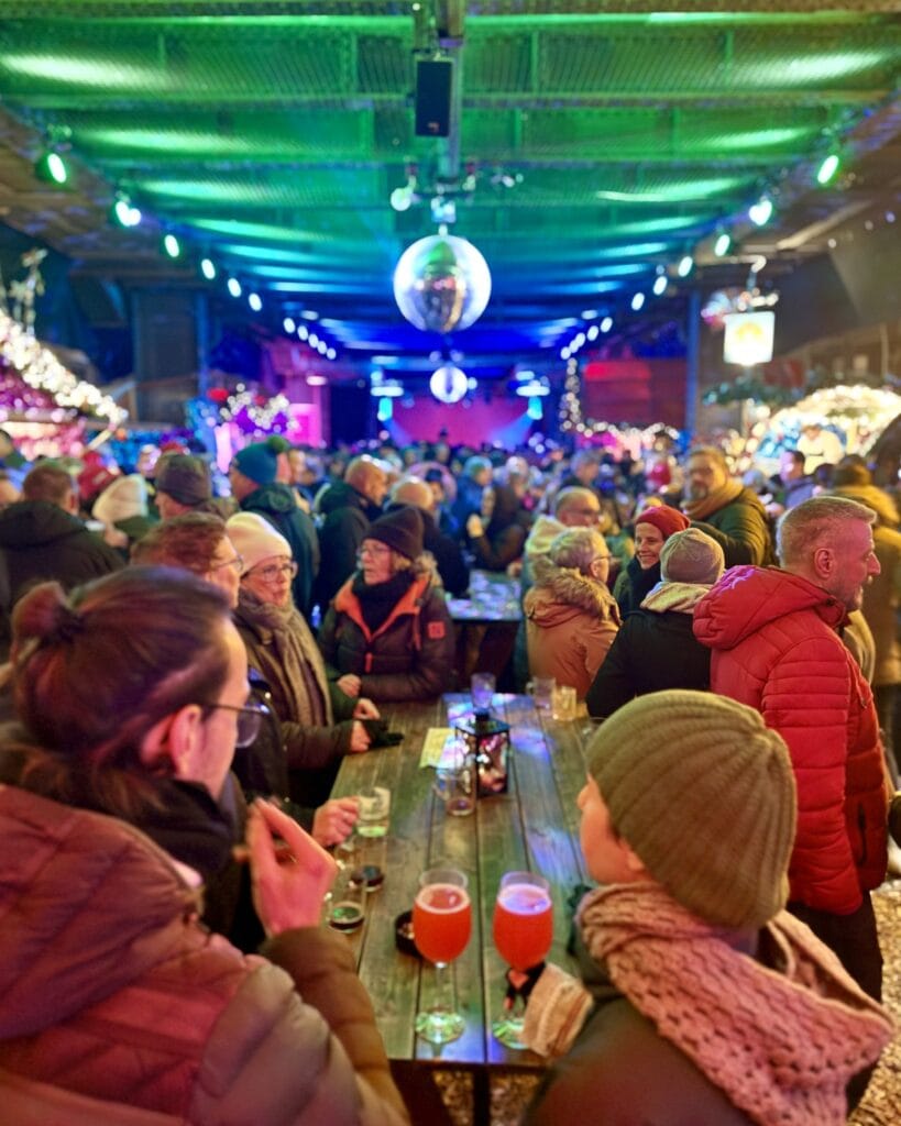 Auf einem Weihnachtsmarkt genießen Besucher an einem langen Holztisch Biere in Gläsern, während im Hintergrund Lichter funkeln.