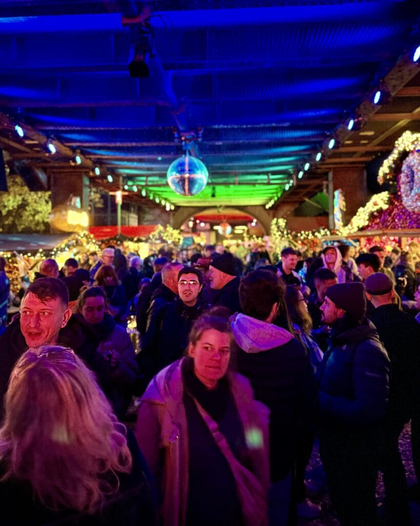 Auf einem Weihnachtsmarkt drängen sich viele Menschen unter einer hell erleuchteten Discokugel und genießen die festliche Atmosphäre.