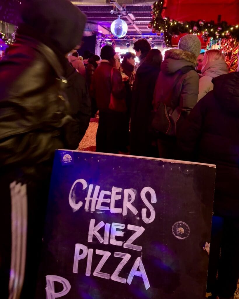 Auf der Christmas Avenue stehen Besucher vor einem Schild, das "Cheers Kiez Pizza" in weißer Schrift anbietet.