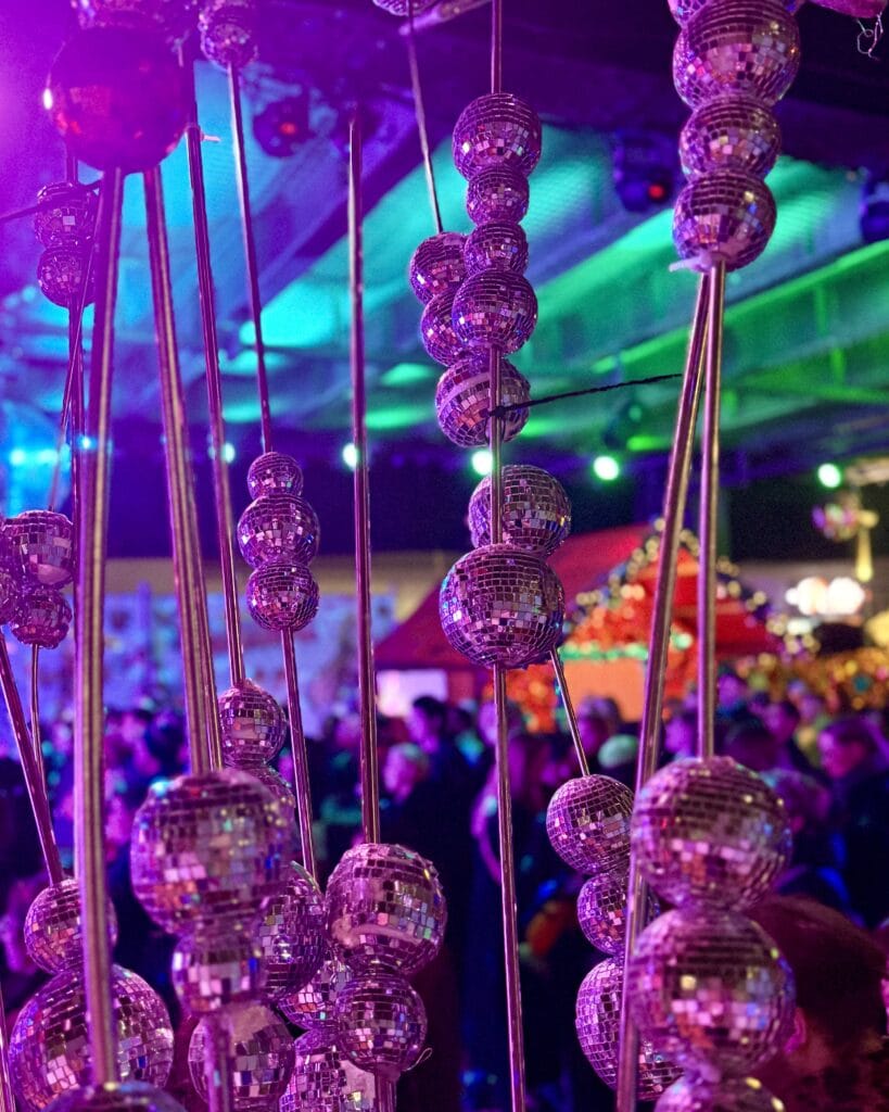 Auf einem Weihnachtsmarkt hängen viele kleine Discokugeln an dünnen Stangen und reflektieren das violette und grüne Licht.