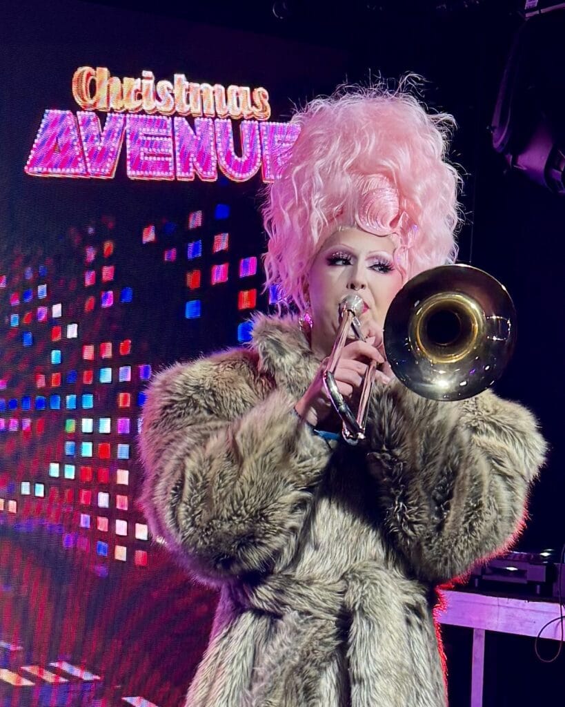 Eine Dragqueen mit rosa Perücke und Fellmantel spielt auf einer Posaune vor dem Schriftzug Christmas Avenue.