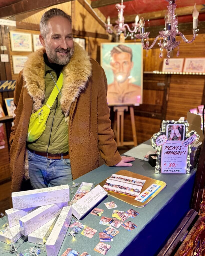 Ein lächelnder Händler präsentiert seine Waren, darunter ein Memory-Spiel für Erwachsene, auf einem Weihnachtsmarkt.