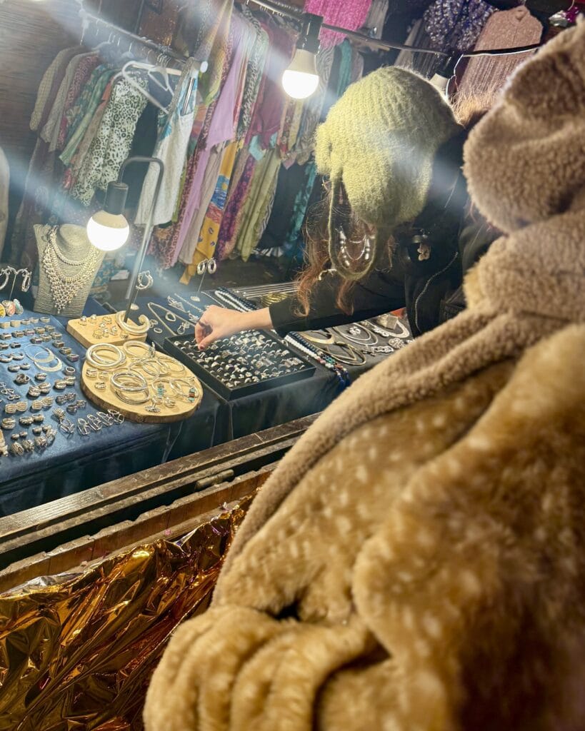 Eine Verkäuferin präsentiert auf einem Markt ihren Schmuck an einem beleuchteten Verkaufsstand mit viel Kleidung im Hintergrund.