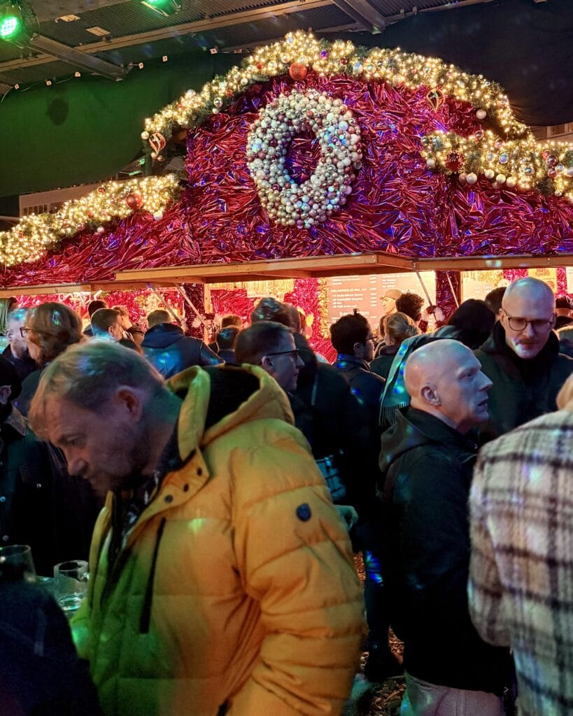 Auf einem Weihnachtsmarkt drängen sich Menschen vor einem dekorierten Stand mit Lichtern und einem großen, runden Weihnachtsschmuck.