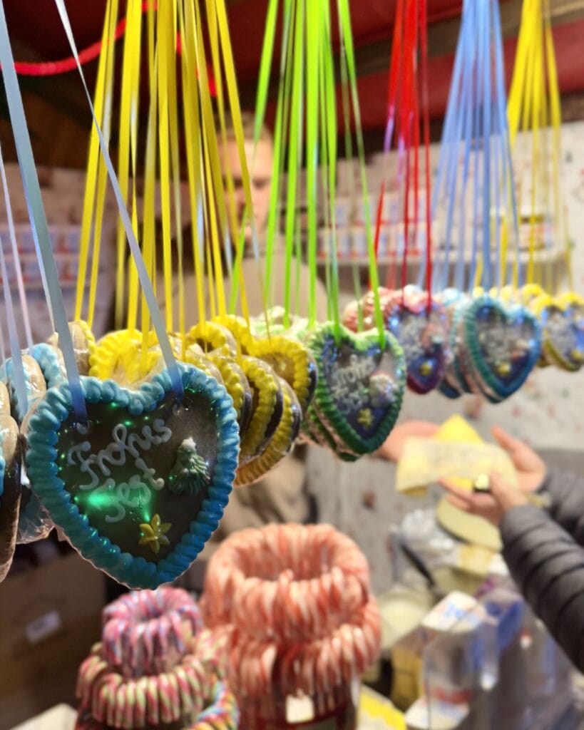Auf einem Weihnachtsmarkt hängen Lebkuchenherzen mit der Aufschrift "Frohes Fest" an farbigen Bändern über einem Verkaufsstand.
