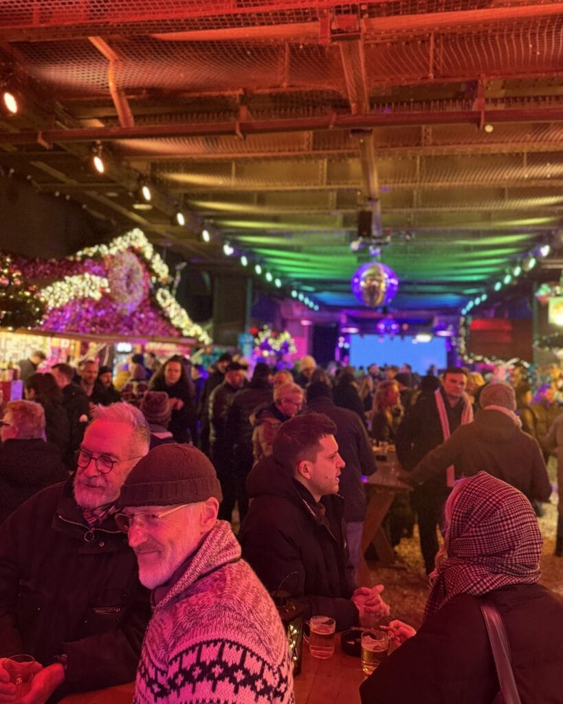 Auf einem Weihnachtsmarkt in Berlin stehen Besucher an einem Tisch und trinken etwas im bunten Licht.
