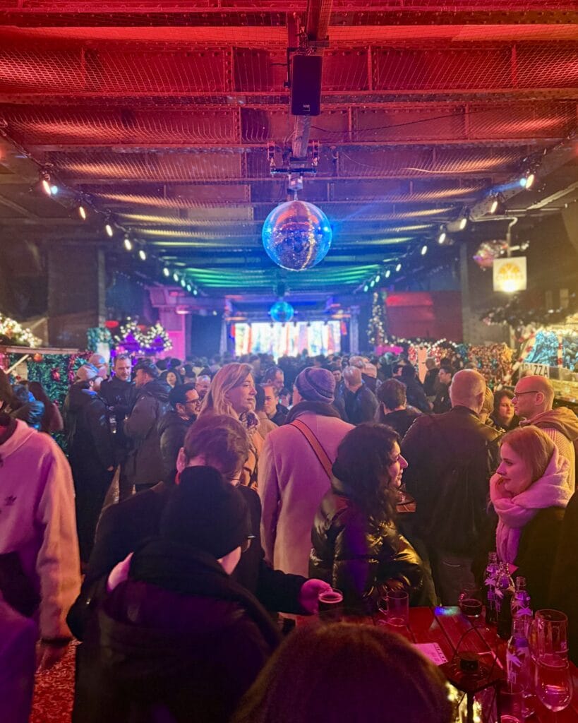 Auf einem Weihnachtsmarkt stehen viele Besucher unter bunten Lichtern und Diskokugeln und unterhalten sich angeregt an einem Stehtisch.