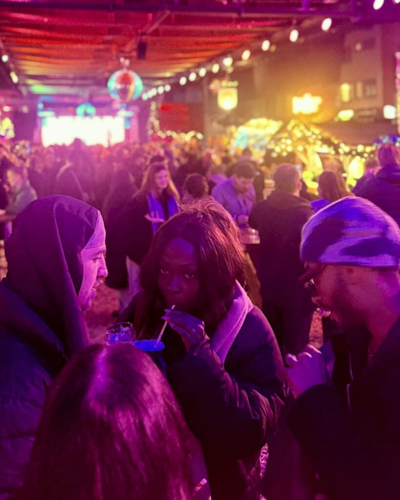 Auf einem Weihnachtsmarkt trinkt eine junge Frau mit einem Strohhalm aus einem blauen Becher, umgeben von anderen Besuchern.