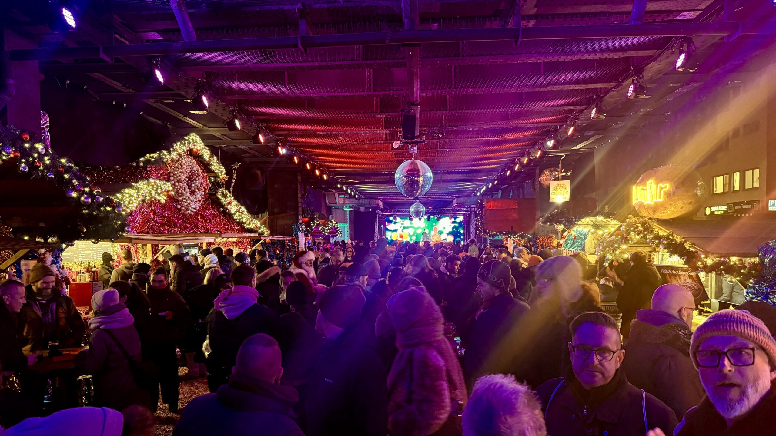 Auf einem Weihnachtsmarkt unter Discokugeln drängen sich viele Menschen vor einer Bühne mit Lichteffekten.