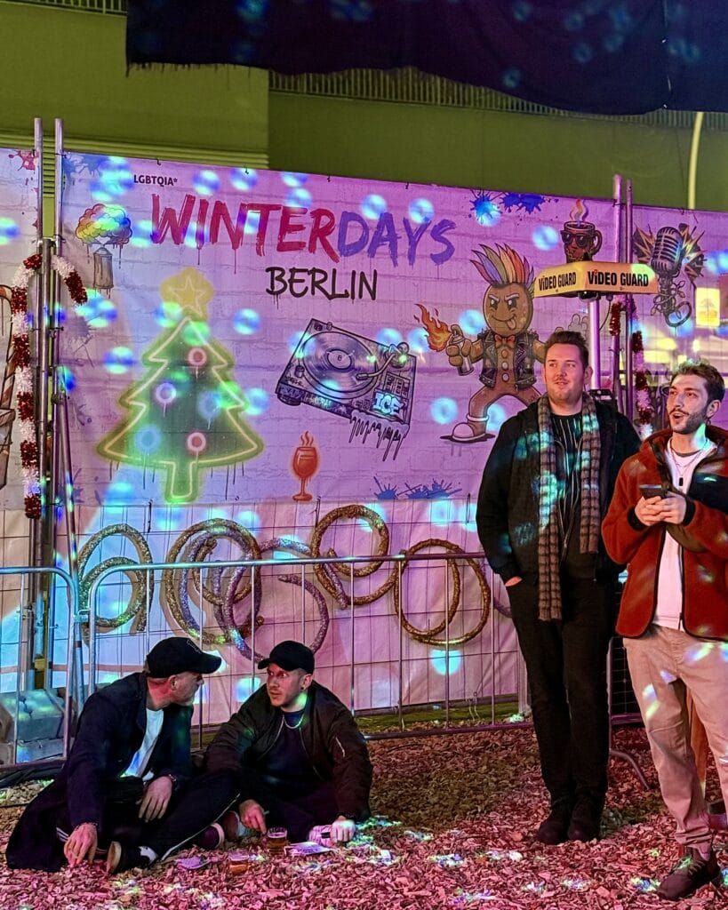 Drei Männer stehen vor einer Wand mit Graffiti und zwei Männer sitzen am Boden und unterhalten sich bei einem Winterevent.