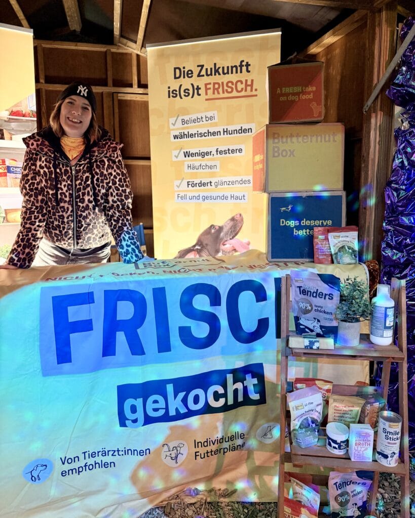 Eine freundliche Frau steht an einem Verkaufsstand für Hundefutter, der mit dem Schriftzug 'FRISCH gekocht' versehen ist.