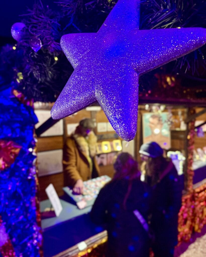 Ein glitzernder, lila Stern hängt über einem Verkaufsstand auf dem Weihnachtsmarkt, wo Besucher einkaufen.
