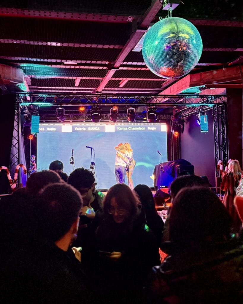 Besucher genießen ein Konzert auf einer Bühne mit Discokugel im Vordergrund, während ein Paar auf dem Bildschirm im Hintergrund zu