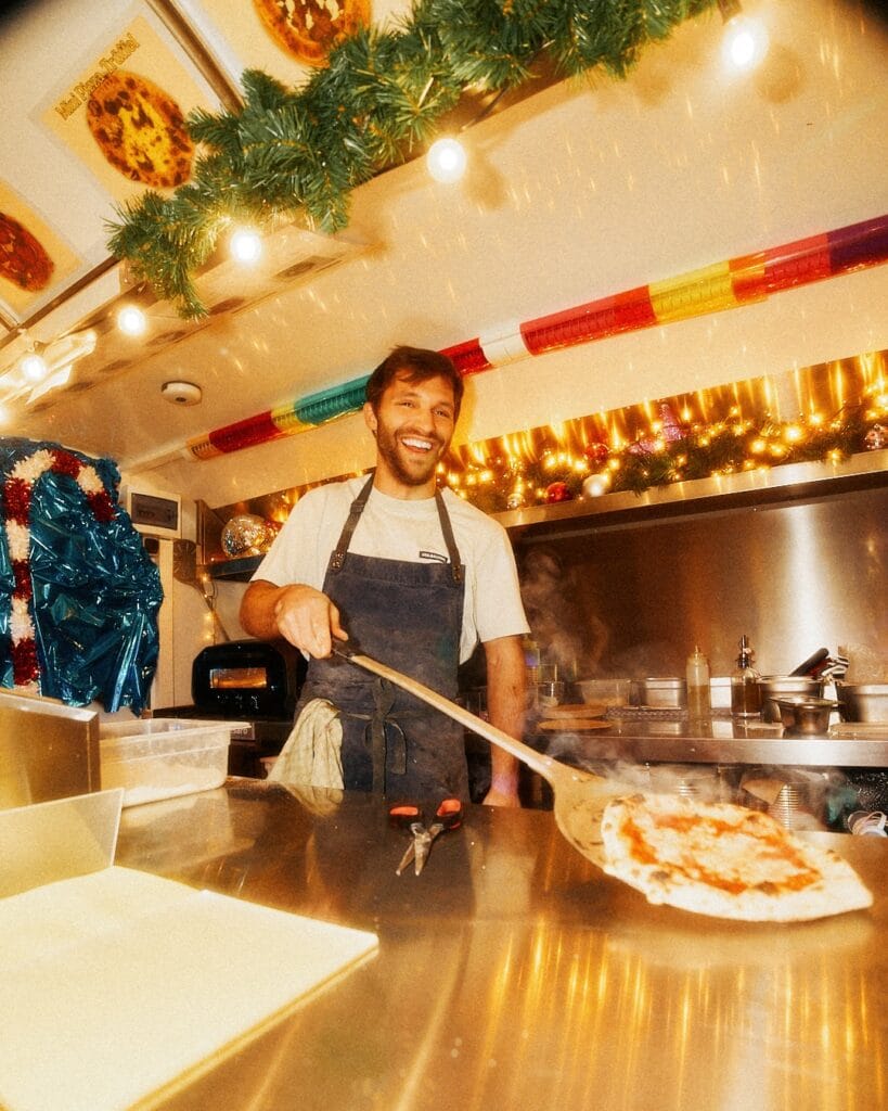 Ein lächelnder Pizzabäcker hantiert mit einer dampfenden Pizza in einem weihnachtlich geschmückten Foodtruck.