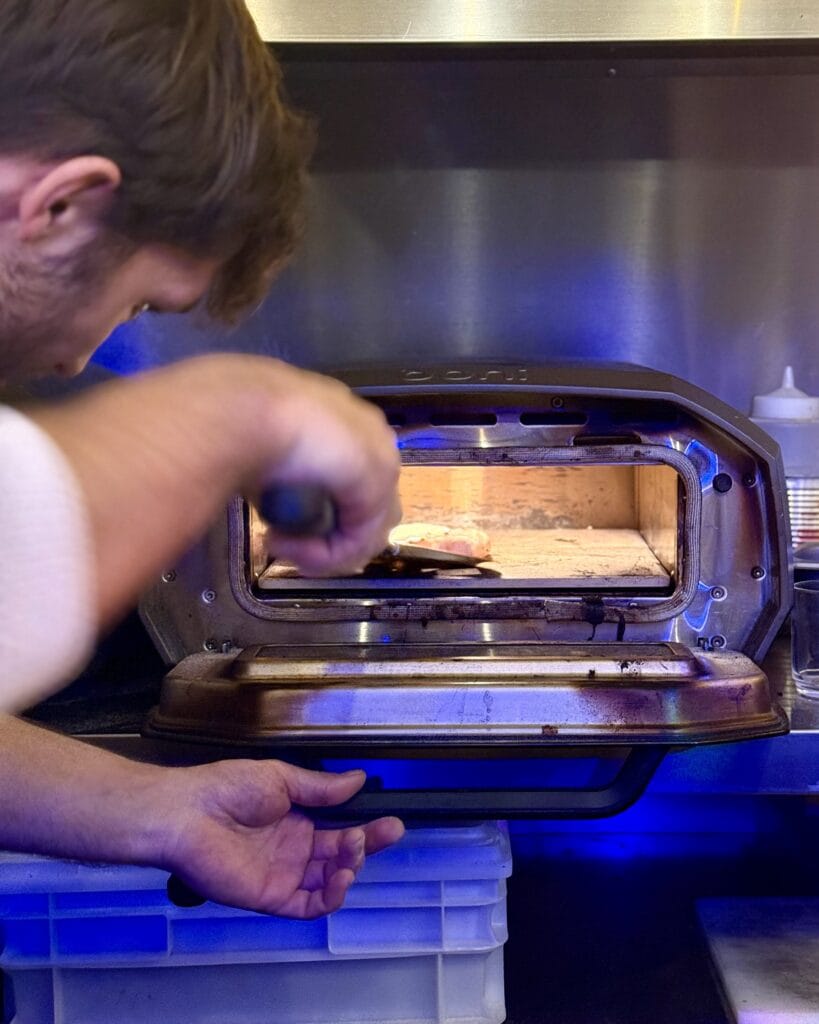 Ein Mann schiebt mit einem Schieber eine Pizza in einen kleinen Pizzaofen. Das Innere des Ofens ist hell erleuchtet.