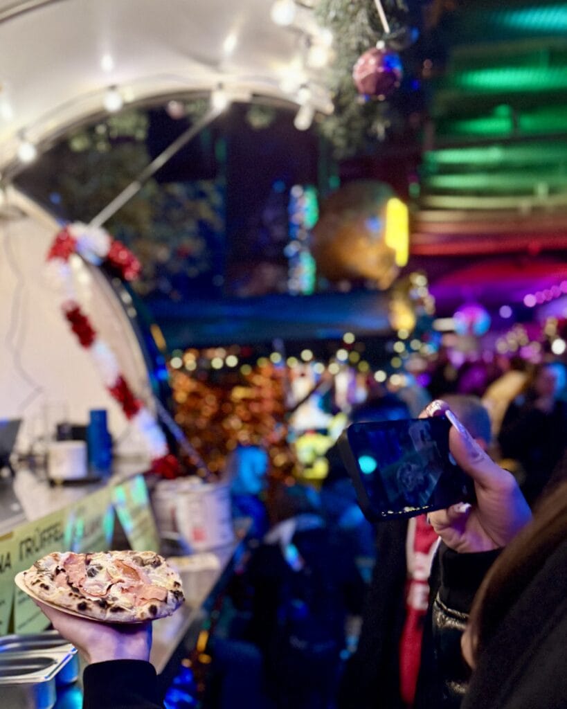 Eine Person hält eine Pizza in der Hand und filmt oder fotografiert sie mit einem Smartphone.