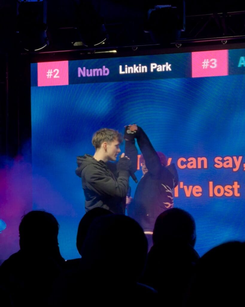 Zwei Sänger performen auf der Bühne vor einer großen Leinwand mit Text und Platzierungen bei einem Karaoke-Event.